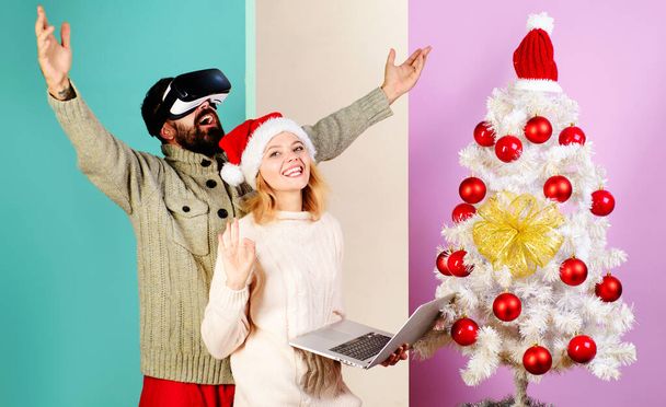 Happy family near Christmas tree. Bearded man in VR glasses and smiling woman in Santa hat with laptop show sign ok. 3d technology. Christmas couple. Winter holiday. Christmas or New Year celebration - Photo, Image