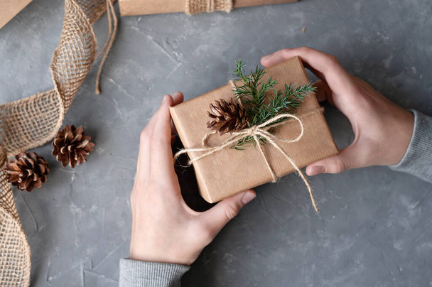 Χέρια ατόμου κρατώντας καφέ δημιουργημένο κουτί δώρου διακοσμημένα με κλαδί πεύκου και κώνο πάνω από γκρι φόντο τραπέζι. Βιώσιμη αισθητική Χριστουγεννιάτικη γιορτή, παρουσιάζει προετοιμασία έννοια. - Φωτογραφία, εικόνα