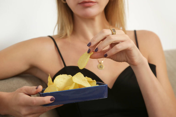 Young sad woman having unhealthy snack, eating potato chips in the livingroom. Junk food and unhealthy eating concept. - Photo, Image
