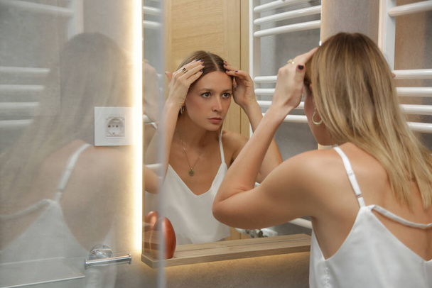 Portrait of a beautiful young woman examining her scalp and hair in front of the mirror, hair roots, color, grey hair, hair loss or dry scalp problem - Photo, Image