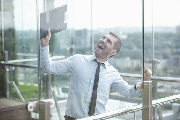Empresario está sosteniendo una carpeta, mirando hacia arriba y gritando de felicidad mientras está de pie en el balcón del edificio de oficinas, vista a través de la ventana
 - Foto, Imagen