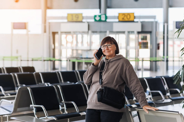 Счастливая улыбающаяся зрелая женщина держит смартфон и машет рукой в приветствии человеку, встретившему ее в терминале аэропорта после прилета из поездки в новую страну на каникулы. Путешествие на пенсию. - Фото, изображение