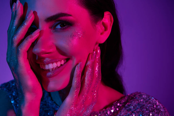 close-up foto van vrolijke dame touch gezicht van toepassing glitter make-up op nieuwjaar themafeest over neon achtergrond. - Foto, afbeelding
