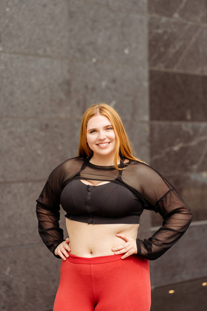 Mujer sonriente usando ropa deportiva y posando contra la pared al aire libre. Cuerpo positivo, además de tamaño, motivación - Foto, Imagen