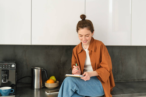 Portré elegáns fiatal nő, ül otthon a konyhában, és jegyzetel, írja le a receptet, ellenőrzi a listát a ház megbízások, firkál vagy rajz notebook. - Fotó, kép