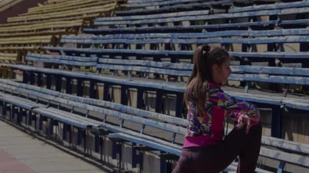 Młoda dziewczyna robi ćwiczenia gimnastyki na stadionie. Rozciągający się na nogi i stopy przed wyścigiem. - Materiał filmowy, wideo