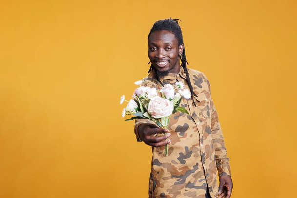 Πορτρέτο του Αφροαμερικανού άνδρα που κρατά λευκό μπουκέτο λουλουδιών, παρουσιάζοντας έκπληξη για τη φίλη του στην κάμερα στο στούντιο πάνω από κίτρινο φόντο. Ρομαντικό αγόρι ποζάρει για την ημέρα του Αγίου Βαλεντίνου - Φωτογραφία, εικόνα