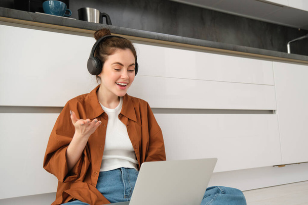 Mujer joven, freelancer se une al chat en línea, estudiante que estudia en casa, se sienta en el suelo con computadora portátil y auriculares. - Foto, imagen
