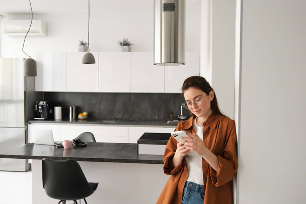Portrait of young woman at home, checking news or messages on smartphone, online shopping, confirm delivery on mobile phone app, leaning on wall, standing in living room. - Photo, Image