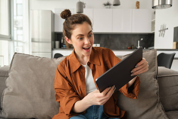 Portrait d'une jeune femme excitée et souriante jouant à des jeux sur sa tablette numérique, penchant gadget et regardant amusé à l'écran, assis sur le canapé à la maison. - Photo, image