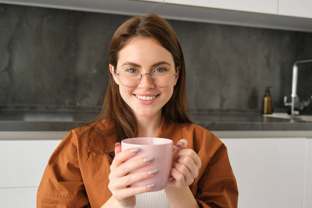 Πορτρέτο της όμορφης νεαρής γυναίκας με γυαλιά, χαμογελαστή, αναπαύεται στο σπίτι, κρατώντας κούπα με καφέ, πίνοντας τσάι. - Φωτογραφία, εικόνα
