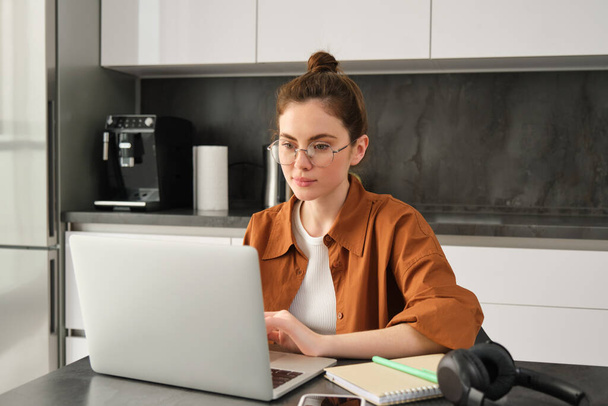 Πορτρέτο της νεαρής συγκεντρωμένης γυναίκας σε γυαλιά, αυτοαπασχολούμενος ελεύθερος επαγγελματίας, που εργάζεται από το σπίτι σε φορητό υπολογιστή, χρησιμοποιώντας υπολογιστή στο σπίτι. - Φωτογραφία, εικόνα