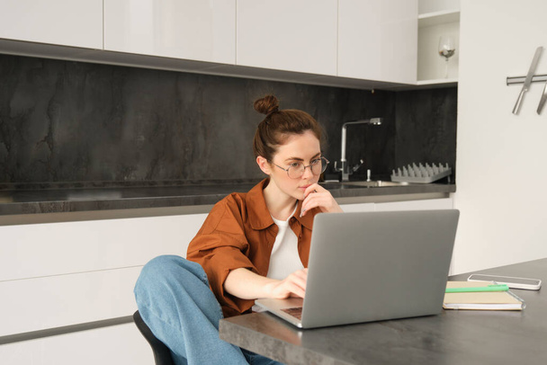 Retrato de mujer sentada en casa con portátil. Empresaria administrando su propio negocio remotamente desde su cocina. Estudiante mirando la pantalla de la computadora, estudiando, haciendo su tarea. - Foto, imagen