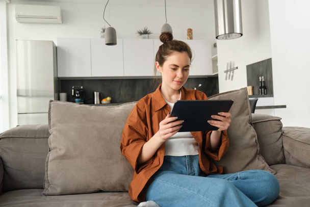 Retrato de una joven sentada en un sofá en la sala de estar, viendo películas en una tableta digital, leyendo en un dispositivo, disfrutando de un fin de semana en casa. - Foto, Imagen