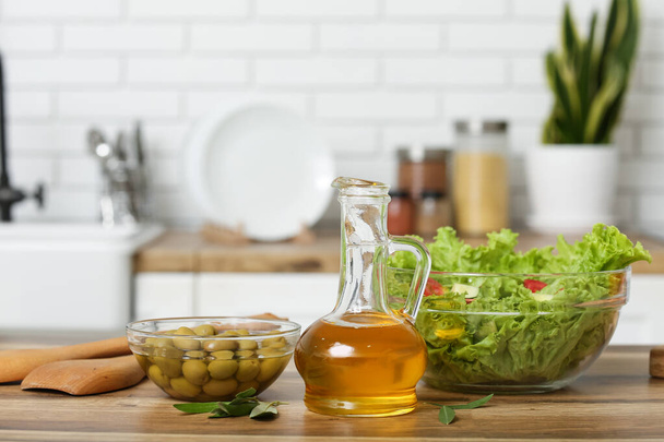 Jarra con aceite, aceitunas y ensalada fresca en la mesa en la cocina - Foto, Imagen
