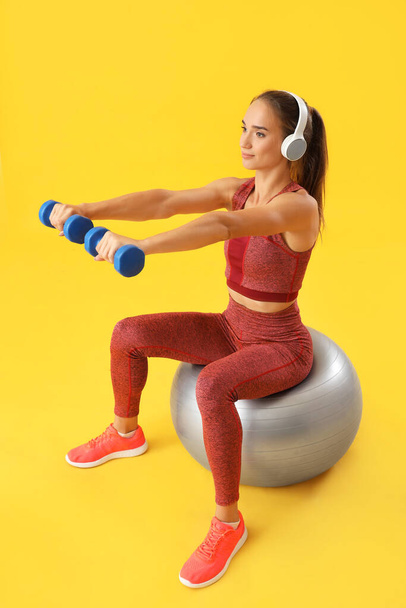 Sportliche junge Frau mit Kurzhanteln und Fitball auf gelbem Hintergrund - Foto, Bild