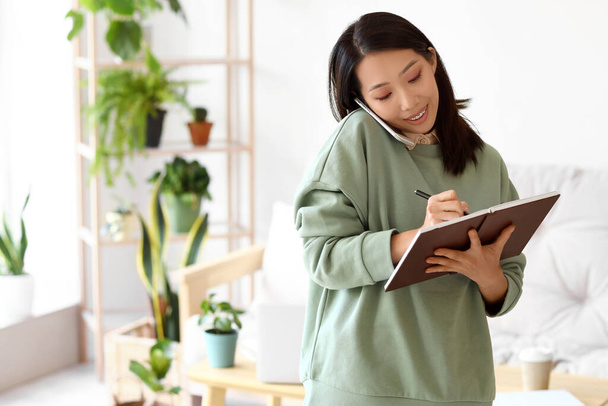 jong aziatische vrouw met notebook praten door mobiele telefoon thuis - Foto, afbeelding