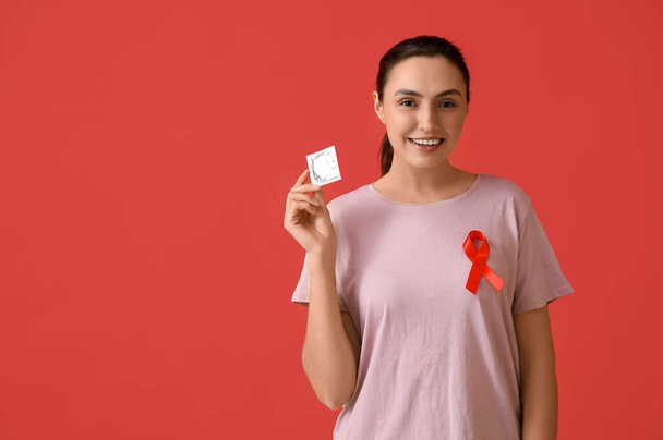 Jonge vrouw met condoom en rood lint op kleur achtergrond. Bewustmakingsconcept - Foto, afbeelding