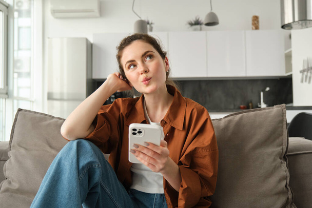 Πορτρέτο της χαριτωμένης περίεργης γυναίκας, κάθεται σε καναπέ με smartphone, κοιτάζει ψηλά και σκέφτεται, δίνοντας μια παραγγελία, online ψώνια σε εφαρμογή για κινητά τηλέφωνα. - Φωτογραφία, εικόνα