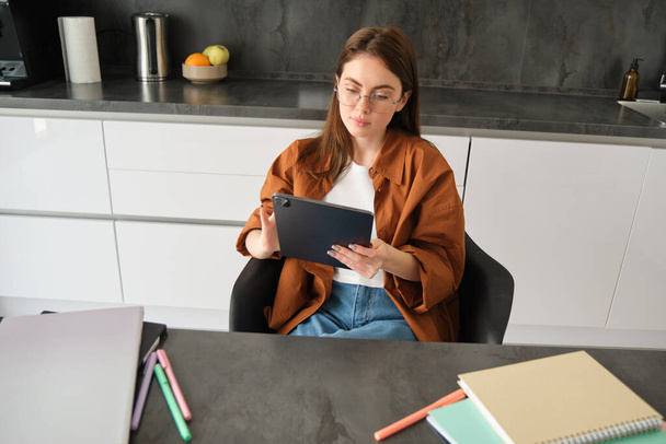 E-öğrenme ve uzaktan eğitim kavramı. Gözlüklü genç bir kadın, mutfakta çalışıyor, dijital tablete bakıyor, online sınıfa ya da kursa bağlanıyor.. - Fotoğraf, Görsel