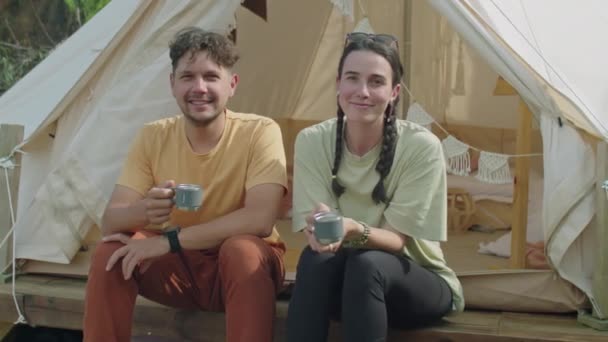 Portrait de jeune couple heureux tenant des tasses à thé, posant à la caméra et souriant tout en étant assis ensemble devant une tente glamping pendant les vacances d'été - Séquence, vidéo