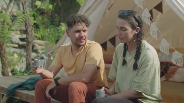 Keskikokoinen kuva nuoresta parista istumassa glamping-teltan ulkopuolella, juomassa teetä ja keskustelemassa ympäröivästä luonnosta viettäessään kesälomaa yhdessä - Materiaali, video