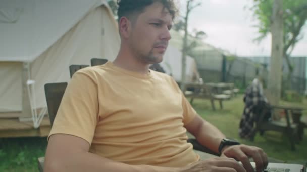 Plan incliné du touriste masculin assis à l'extérieur devant la tente sur le glampsite, tapant sur un ordinateur portable et regardant la nature pendant les vacances - Séquence, vidéo