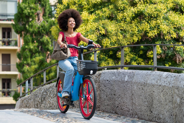 Elinde çantasıyla gülümseyen Faslı genç bir kadının şehir köprüsünde elektrikli bisiklet sürerken gün ışığında bulanık yeşil ağaçlara ve binaya sürüşünün yan görüntüsü. - Fotoğraf, Görsel