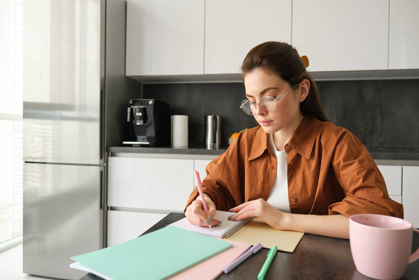 Retrato de mujer mirando concentrada, anotando notas, haciendo deberes en la cocina, tomando café, estudiando en casa. - Foto, Imagen