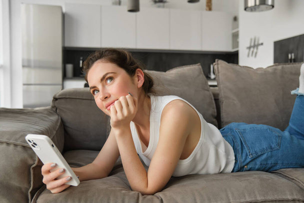 İnternetten alışveriş konsepti. Kanepede uzanan genç bir kadın, akıllı telefonuyla kanepede uzanıyor, yukarı bakıyor ve düşünüyor.. - Fotoğraf, Görsel