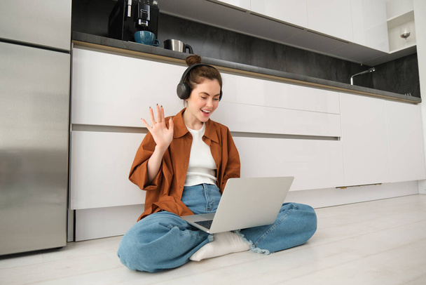 Retrato de la mujer feliz en los auriculares, mirando a la computadora portátil, saludando la mano en la computadora, charlando en video en línea, se une a la reunión del equipo, llamando a un amigo en la computadora, sentado en el suelo. - Foto, Imagen