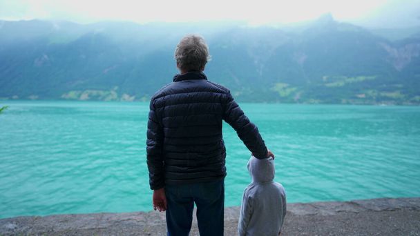 Tył dziadka i wnuka stojących przed jeziorem i widokiem na góry. Rodzina cieszy się na świeżym powietrzu w kurtki w deszczu mżawki, spokojna i spokojna scena - Zdjęcie, obraz