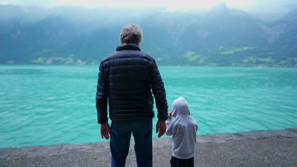 湖と山の景色の前に立っている祖父と孫の背中. 乾燥した雨の中のジャケットを着た素晴らしいアウトドアを楽しむ家族,穏やかで平和なシーン - 写真・画像