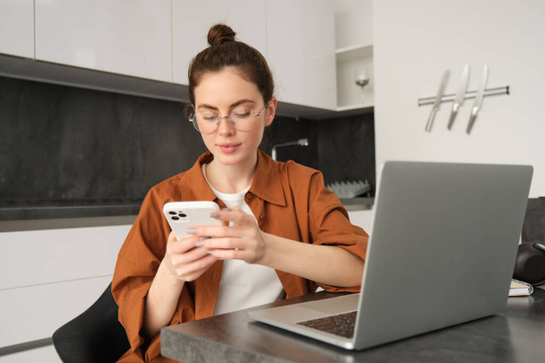 Retrato de jovem freelancer feminino, mulher trabalhando em casa com laptop, programadora sentada com computador na cozinha, mensagem de resposta no smartphone. - Foto, Imagem