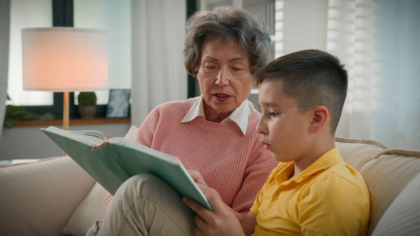 Любляча літня жінка на пенсії, бабуся читає книгу з онуком вдома на дивані, насолоджуючись читанням цікавої казкової історії, піклуючись про бабусю, допомагаючи дитині хлопчику з домашнім завданням, вивчаючи енциклопедію - Фото, зображення