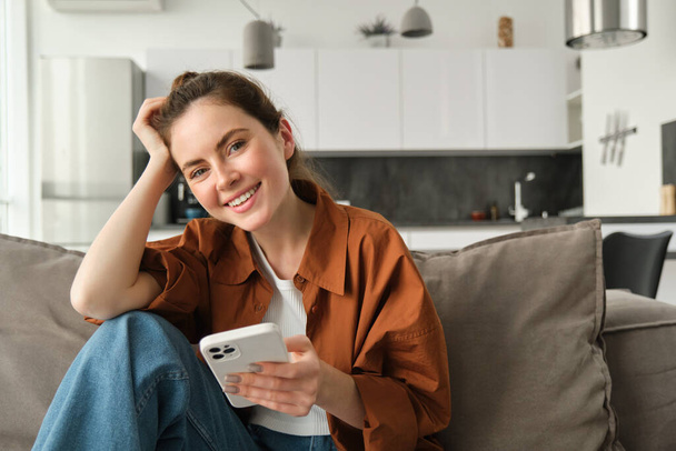 Portrait of cute young smiling woman, sitting on sofa with mobile phone, relaxing at home and using smartphone, scrolling social media, buying online, ordering something on application. - Photo, Image