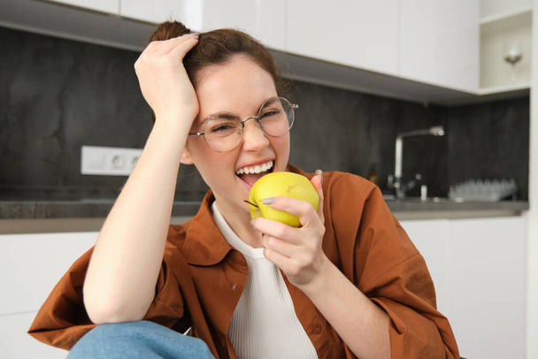 Ritratto ravvicinato di giovane donna carina in occhiali, mangiare mela, ridere e sorridere, seduta in cucina. - Foto, immagini