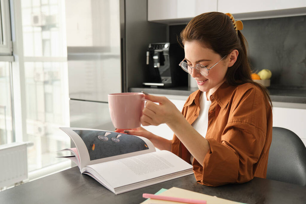 Πορτρέτο της γυναίκας που διαβάζει στο σπίτι, γυρίζοντας σελίδες του αγαπημένου βιβλίου, χαλαρώνοντας στην κουζίνα με φλιτζάνι καφέ, φορώντας γυαλιά. - Φωτογραφία, εικόνα