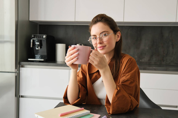 Portrait of beautiful brunette woman, sitting in kitchen, taking a break for cup of coffee. Girl drinks tea at home. - Foto, Imagen