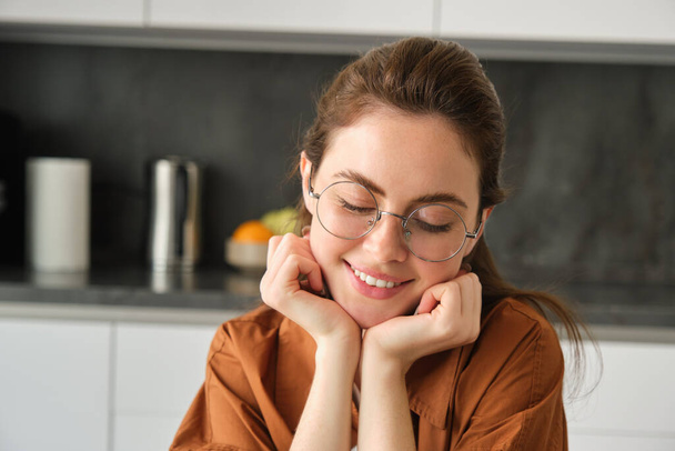 Porträt einer niedlichen, schönen jungen Frau mit Brille, die zu Hause in der Küche sitzt, Kopf an Hände lehnt, verträumt lächelt und blickt. - Foto, Bild