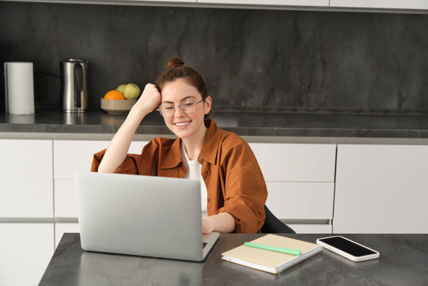 Πορτρέτο της γυναίκας ελεύθερος επαγγελματίας, προγραμματιστής που εργάζονται από το σπίτι. Γυναίκα φοιτητής ή ιδιοκτήτης επιχείρησης κάθεται με φορητό υπολογιστή στην κουζίνα. - Φωτογραφία, εικόνα