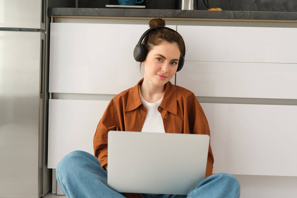 Νεαρή γυναίκα, μαθήτρια που κάνει την εργασία στο φορητό υπολογιστή, κάθεται στο πάτωμα, ακούγοντας μουσική στα ακουστικά και εργάζεται από το σπίτι. - Φωτογραφία, εικόνα