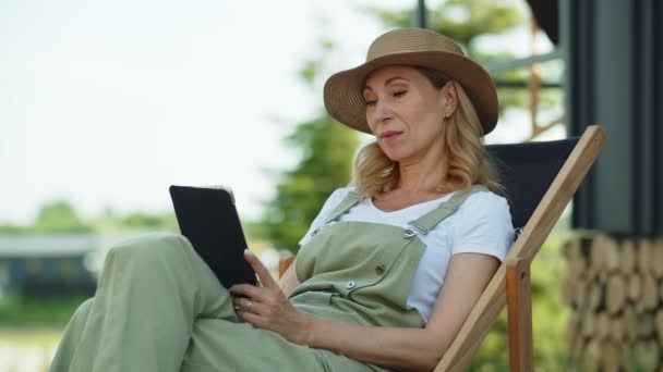 Žena středního věku tráví letní dovolenou na venkově, užívá si venku a odpočívá po práci na zahradě, sedí na odpočívadle. Vážná dáma používá tablet a čte si eBook s hlubokým - Záběry, video