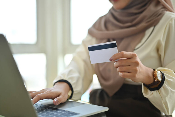 Μουσουλμάνα γυναίκα που κατέχει πιστωτική κάρτα χρησιμοποιώντας φορητό υπολογιστή κάνει online τραπεζική συναλλαγή ή ψώνια στο ηλεκτρονικό κατάστημα.. - Φωτογραφία, εικόνα