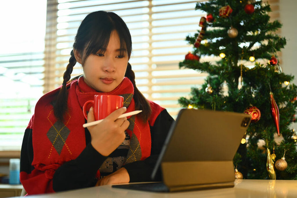 Hübsche junge Frau im warmen Pullover trinkt Kaffee und liest E-Mails auf dem digitalen Tablet. - Foto, Bild