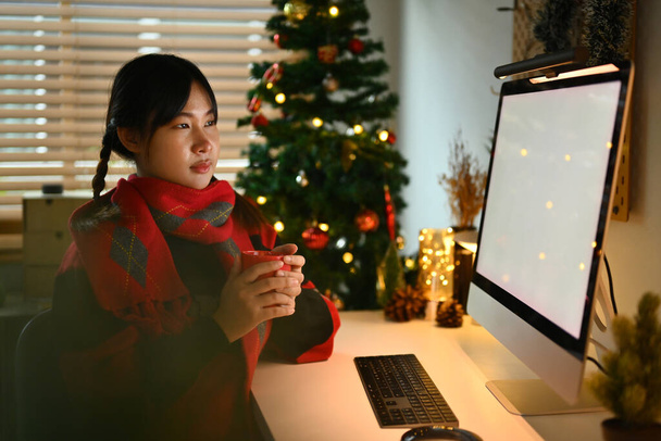 Junge Frau hält Becher mit heißer Schokolade in der Hand und blickt auf Computermonitor mit Weihnachtsbaum im Hintergrund. - Foto, Bild