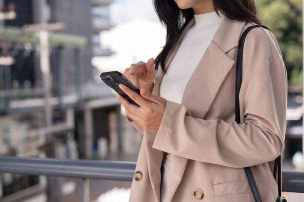 Zugeschnittene Seitenansicht einer schönen asiatischen Geschäftsfrau in einem trendigen Businessanzug, die ihr Smartphone benutzt, E-Mails checkt oder auf Nachrichten reagiert, während sie auf dem Skywalk spaziert. - Foto, Bild