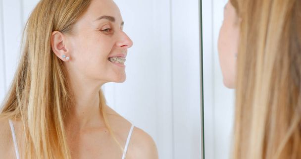 Молодая женщина, щекочущая свои новые брекеты в зеркале, зубные брекеты концепции гигиены полости рта. Высокое качество фото - Фото, изображение