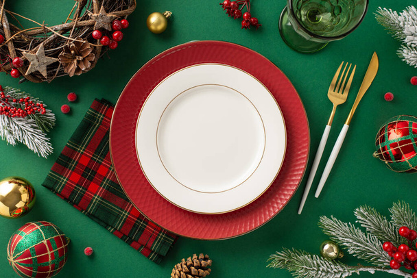 Elegant holiday table setup idea. Overhead shot showcasing dishes, golden utensils, plaid napkin, wine goblet, ornaments, rustic wreath, icy fir branches, mistletoe on a green backdrop - Photo, Image
