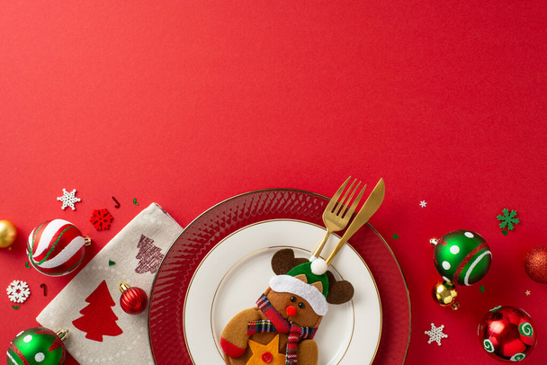 Festive kids' table setup for New Year's party. Overhead shot of plates, cute reindeer-shaped utensil holder, festive napkin, baubles, snowflake confetti on a red backdrop - Photo, Image